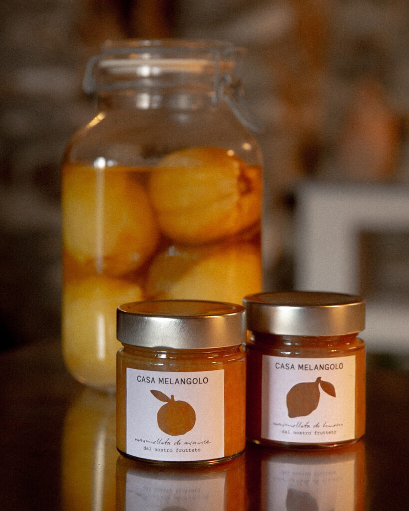 This image showcases artisanal food products from "Casa Melangolo," featuring two jars of marmalade in the foreground and a large jar of preserved lemons in the background. The marmalade jars, each with a golden lid, display minimalist labels. One jar is labeled "Marmellata di arance" (orange marmalade), featuring the silhouette of an orange cut out from the label, while the other is labeled "Marmellata di limoni" (lemon marmalade). Both labels mention "dal nostro frutteto" (from our orchard), highlighting the homemade, farm-to-table nature of the products. The jar of preserved lemons behind them adds to the rustic, handcrafted aesthetic of the scene.