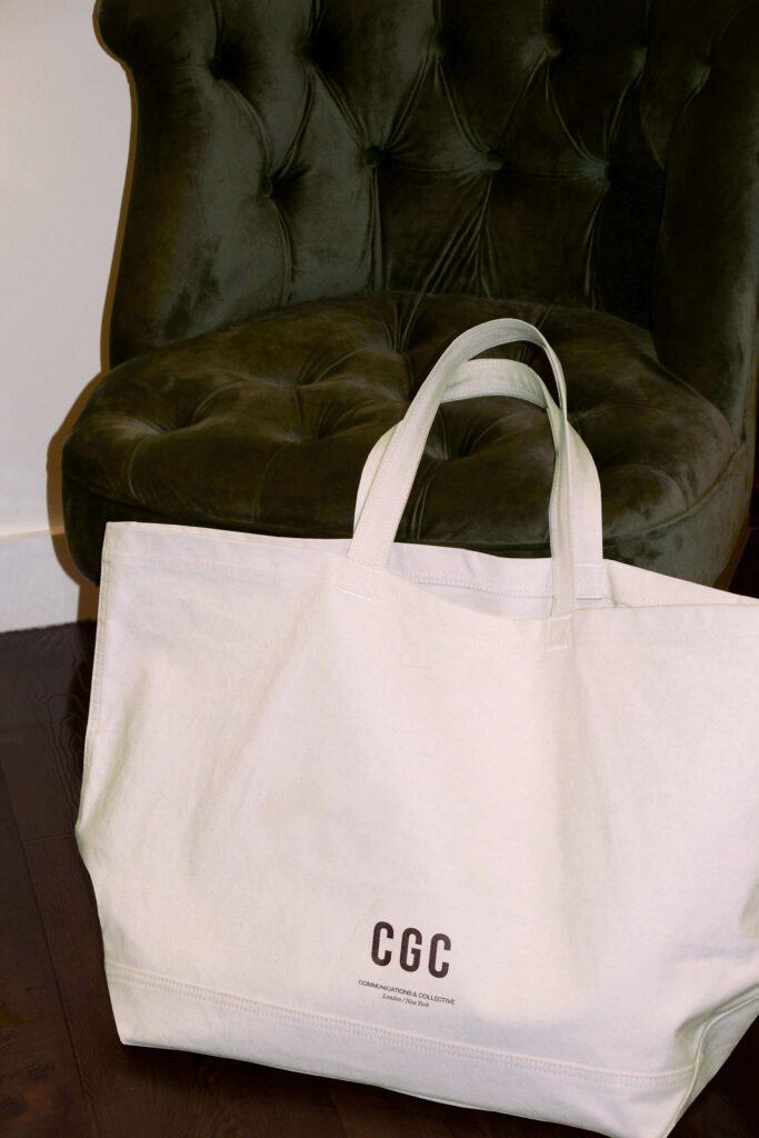 A photograph of a white tote bag on a green velvet sofa with Clare Goodwin Communications & Collective's black logo printed in black.